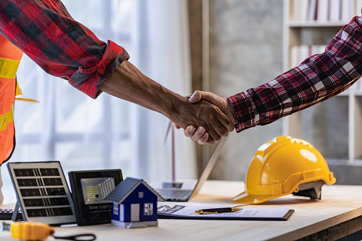 Yellow hard hat on table and house model solar windmill with construction team handshake to start new project contract plan in renewable energy concept contractor office center
